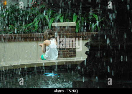 Vista attraverso le gocce della fontana sulla piccola ragazza seduta da sola nel giardino tropicale. Triste kid è in attesa di qualcuno. Concetto di adozione Foto Stock