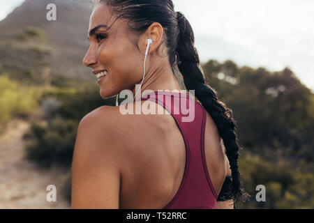 Vista posteriore di una femmina di runner in piedi su una strada sterrata in mattinata con gli auricolari che guarda lontano e sorridente. Donna in attrezzature fitness pronto per il cross country Foto Stock