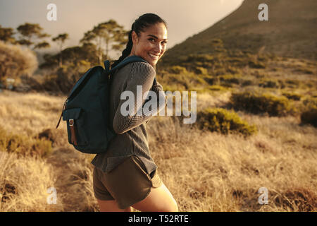 Donna con zaino a piedi fino al paese trail e guardando sopra la sua spalla e sorridente. Foto Stock