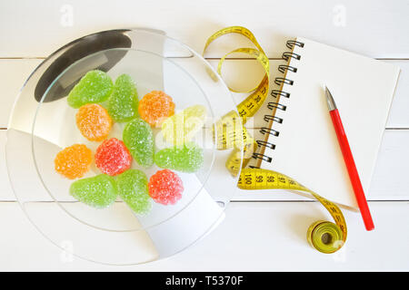 Gelatine di frutta sulla bilancia da cucina accanto a un notebook, una penna e un metro a nastro. Il conteggio e la registrazione della quantità di proteina, calorie e grassi negli alimenti. Sl Foto Stock