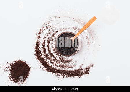 Cosmetici macchia di caffè in barattolo con il cucchiaio di legno accanto alla manciata di ingredienti: grani di terra e lo zucchero sul tavolo bianco, vista dall'alto. Foto Stock