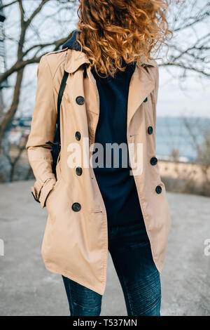 Irriconoscibile giovane donna che indossa il beige trench coat con pulsanti in piedi nel parco della città tenendo le mani nelle tasche, i suoi capelli che fluttua nel vento. Foto Stock