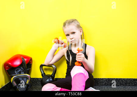 Tema sport e salute dei bambini. Poco divertente per bambini ragazza caucasica con pig-tail, siede pausa di riposo sul pavimento in palestra. Atleta dumbbell attrezzature per Foto Stock