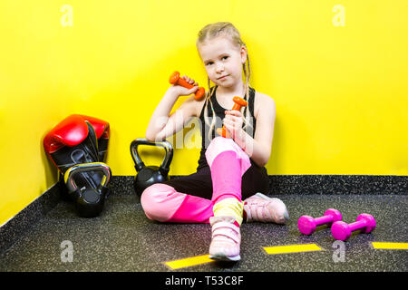 Tema sport e salute dei bambini. Poco divertente per bambini ragazza caucasica con pig-tail, siede pausa di riposo sul pavimento in palestra. Atleta dumbbell attrezzature per Foto Stock