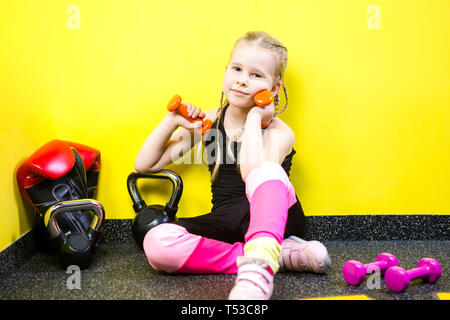 Tema sport e salute dei bambini. Poco divertente per bambini ragazza caucasica con pig-tail, siede pausa di riposo sul pavimento in palestra. Atleta dumbbell attrezzature per Foto Stock