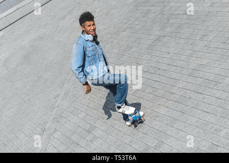Giovane mulatto uomo che indossa le cuffie intorno al collo di skateboard equitazione sulla strada di città cercando fotocamera a sorridere giocoso vista superiore Foto Stock