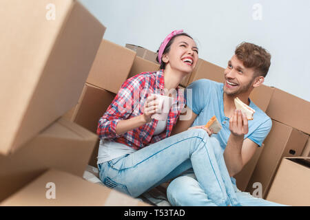 Giovane marito e moglie spostando di nuovo posto seduto sul pavimento ridendo a scherzo loud gioiosa azienda panini e tazze di tè caldo Foto Stock