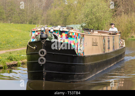 Paesaggio primo piano di una barca stretta in arrivo che viaggia sul canale britannico sotto il sole primaverile; uomo isolato e in pensione in cappellino si trova sul canale posteriore della barca. Foto Stock