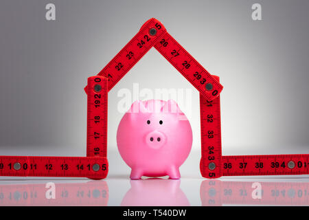 Una vista in elevazione del piccolo rosso Piggybank sotto la Casa fatta con nastro di misurazione Foto Stock