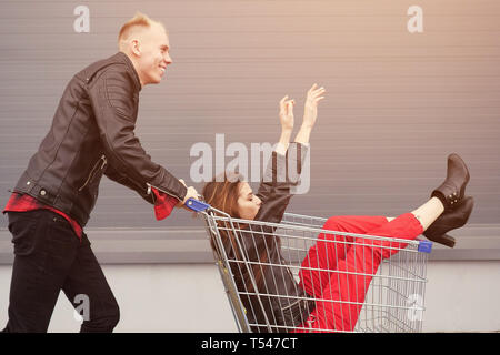 Guy rotoli la sua attraente giovane ragazza nel carrello sul parcheggio del supermercato, alla luce del sole Foto Stock