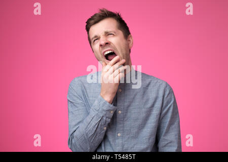 Stanco uomo sente assonnato, sbadigli come si sente stanco, apre la bocca ampiamente, pone su sfondo rosa. Foto Stock