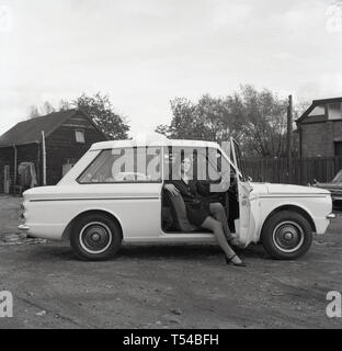 1967, storico, giovane donna che indossa un capo di lana cardigan e gonna seduto nel sedile del guidatore di un Sunbeam Sport autovettura, la versione sportiva della famosa piccola auto, HIllman Imp. Hillman Imp è stata una piccola autovettura realizzato dal gruppo Rootes e il suo successore Chrysler Europa dal 1963 fino al 1976 è stato il concorrente nella piccola auto categoria per il Mini. Foto Stock