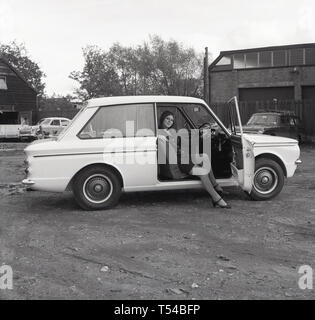 1967, storico, giovane donna che indossa un capo di lana cardigan e gonna seduto nel sedile del guidatore di un Sunbeam Sport autovettura, la versione sportiva della famosa piccola auto, HIllman Imp. Hillman Imp è stata una piccola autovettura realizzato dal gruppo Rootes e il suo successore Chrysler Europa dal 1963 fino al 1976 è stato il concorrente nella piccola auto categoria per il Mini. Foto Stock