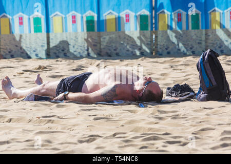 Bournemouth Dorset, Regno Unito. 21 apr 2019. Regno Unito: meteo canicola continua con caldo e soleggiato, come testa beachgoers al mare per godere del calore e del sole a Bournemouth spiagge per le vacanze di Pasqua - metà mattina e già le spiagge sono sempre imballati, come sunseekers arrivarci presto per ottenere il loro spazio. L uomo a prendere il sole da frangivento con cabine sulla spiaggia, design. Credito: Carolyn Jenkins/Alamy Live News Foto Stock