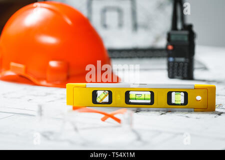 Misuratore di livello con arancione sfocata elmetto, laptop con disegni e walkie talkie su uno sfondo. Architettura e costruzione concept Foto Stock