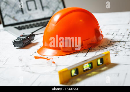 Orange elmetto, laptop con disegni, bicchieri e walkie-talkie con blueprint su un tavolo. Architettura e costruzione concept Foto Stock