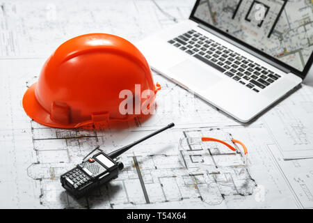 Orange elmetto, laptop con disegni, bicchieri e walkie-talkie con blueprint su un tavolo. Architettura e costruzione concept Foto Stock