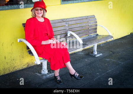 TAURANGA NUOVA ZELANDA; aprile; 20 2019; una donna in abito rosso e cappello seduto da solo dalla parete di colore giallo. Foto Stock