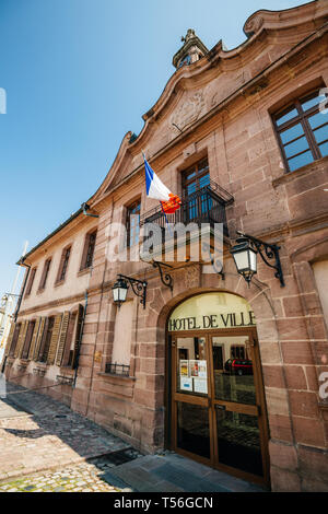 Bergheim, Francia - 19 Apr 2019: Hotel de Ville municipio nella piazza centrale di un ampio angolo di visione con la bandiera francese sventola sopra la porta di ingresso Foto Stock