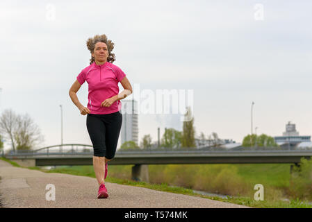 La mezza età della donna in rosa training shirt correndo all'aperto lungo la passerella nel parco. A tutta lunghezza angolo basso vista frontale Foto Stock