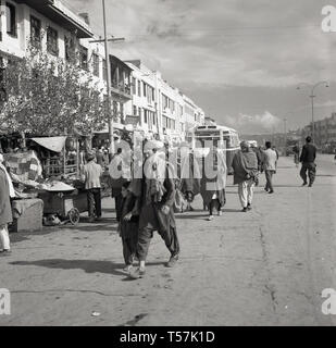 Degli anni Cinquanta, Kabul, Afghanistan, locali afghani in abito nazionale in una ampia strada polverosa passando le bancarelle del mercato nel centro della citta'. Foto Stock