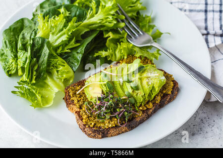 Avocado toast con pesto, germogli e insalata su una piastra bianca. Foto Stock