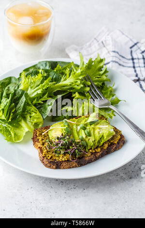 Avocado toast con pesto, germogli e insalata su una piastra bianca e caffè ghiacciato. Foto Stock