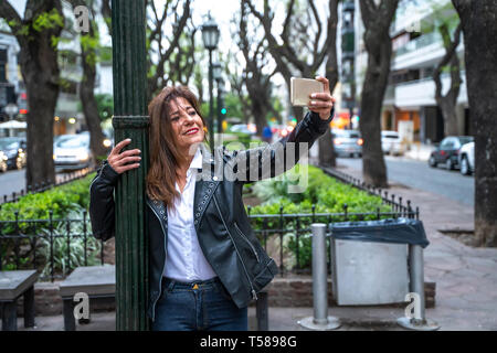 Un sorridente donna matura in un nero giacca di pelle in piedi accanto ad un lampione e prendendo un selfie con il suo smartphone. Foto Stock