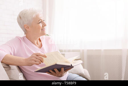 Sognante donna senior in un momento di relax a casa con il libro preferito Foto Stock