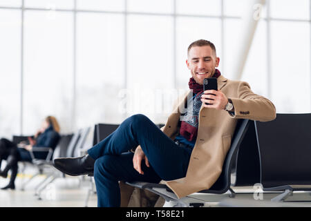 Attraente giovane uomo adulto in Elegante sciarpa e rivestire seduti all'interno di terminal aeroporto area di attesa. Egli guardando di telefonia mobile cellulare e schermo Foto Stock