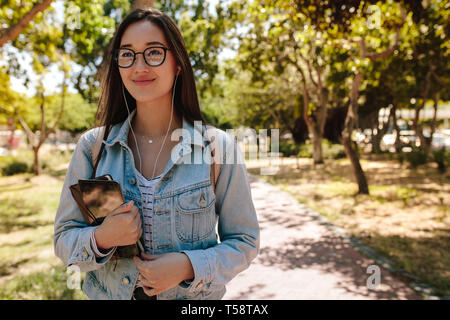 Ritratto di una giovane studentessa camminando nel campus tenendo un tablet pc e prenota. Sorridente studente asiatico a camminare in un parco e ascolto di musica. Foto Stock