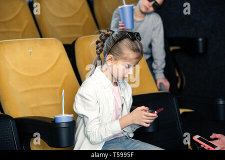 Messa a fuoco selettiva di carino bambino utilizza lo smartphone mentre spendere tempo nel cinema insieme con gli amici Foto Stock