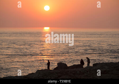 Persone in piedi sulla spiaggia rocciosa di Portland Bill vicino alla famosa attrazione di pulpito Rock sulla punta dell'isola di Portland come il sole va d Foto Stock