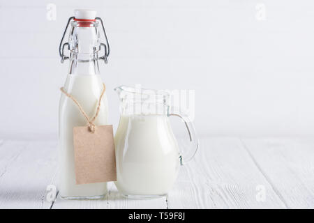 Bottiglia di latte con carta bianca tag sulla fune e la brocca del latte nel dipinto di bianco di tavolo in legno ancora vita Foto Stock