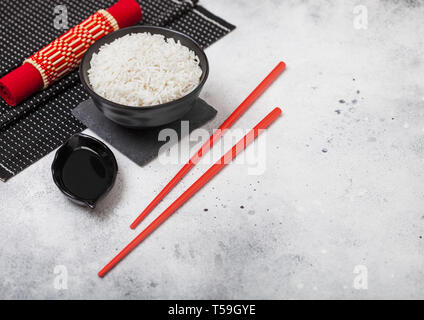 Tazza nera con bolliti basmati organico di riso al gelsomino con bacchette rosso dolce e salsa di soia sul cartellone di bambù con red biancheria sulla cucina luce t Foto Stock