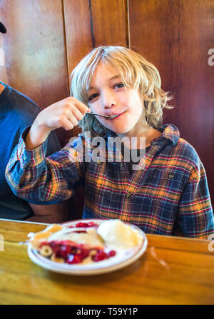 Un giovane ragazzo in un flanel shirt mangia un morso di ciliegia torta in una tavola di legno di Foto Stock