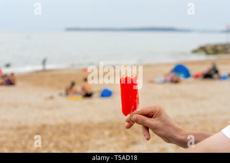 Uomo che tiene una ghiacciola rossa in mano su una spiaggia nel Regno Unito durante un'ondata di caldo in estate, Inghilterra Foto Stock