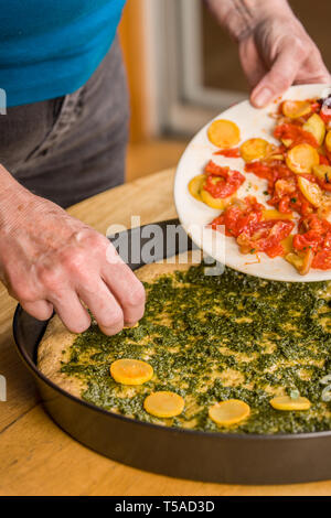 Donna immissione saltata di estate squash sulla sommità del suo in casa multi-grain pizza al pesto, pomodori e arrostito peperoni rossi. (MR) Foto Stock