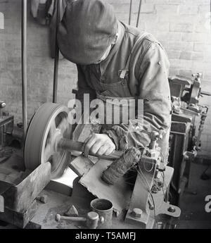 Degli anni Cinquanta, storico, un operaio che indossa il vecchio logori vestiti e un panno il tappo usando una circolare per la macinazione meccanica o con levigatrice su un banco di lavoro in una piccola unità industriali o di laboratorio, Inghilterra, Regno Unito. In seguito tali smerigliatrici da banco sarebbe venuto con un coperchio di protezione o protezione. Foto Stock