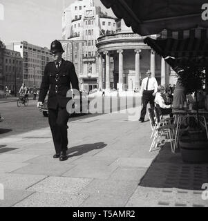 Anni sessanta, storico, Marylebone, London, all'estremità nord di Regent Street, un poliziotto britannico in proprio e a piedi camminando lungo un marciapiede. BBC Broadcasting House in Portland Place e tutte le anime chiesa in Langham Place anche nella foto a distanza. Foto Stock