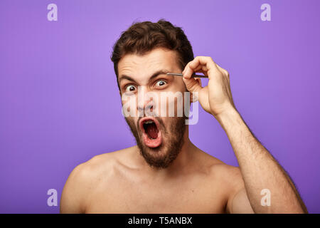 Emotional Man con la bocca spalancata spenna sue sopracciglia con pinzette in mattinata. pelle, cura del viso, il concetto di bellezza passatempo. Foto Stock