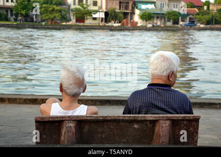 Vista posteriore di due uomini anziani seduti su una panchina che guarda ad un lago in Ha Tinh, Vietnam, nel maggio 2017. Foto Stock