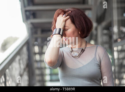 Closeup ritratto di attraente femmina senior mantenendo gli occhi chiusi tenendo la mano sulla testa con una faccina triste espressione isolato su un sfocata background urbano w Foto Stock