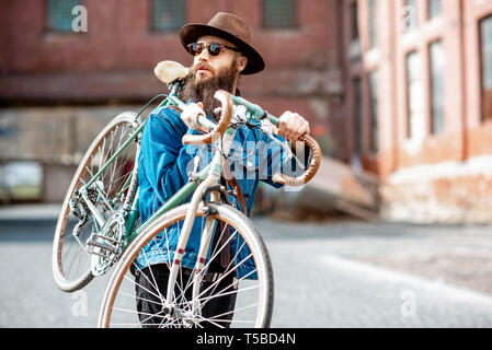 Stile di vita ritratto di un barbuto hipster vestito elegantemente con il cappello e giacca portando la sua bicicletta retrò sul background urbano Foto Stock