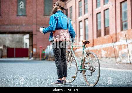 Tanga elegante a piedi con caffè e bicicletta retrò sul industriale di background urbano, vista posteriore Foto Stock