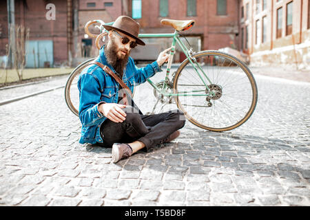 Ritratto di un barbuto hipster vestito elegantemente con il cappello e giacca seduta con retro bicicletta su strada sul background urbano Foto Stock