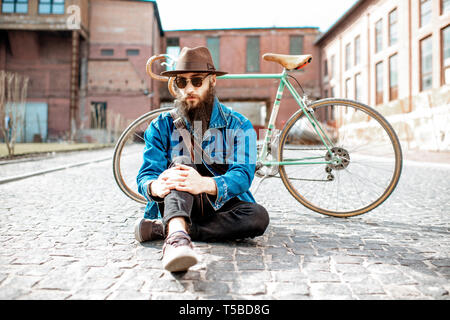 Ritratto di un barbuto hipster vestito elegantemente con il cappello e giacca seduta con retro bicicletta su strada sul background urbano Foto Stock