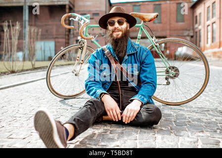 Ritratto di un barbuto hipster vestito elegantemente con il cappello e giacca seduta con retro bicicletta su strada sul background urbano Foto Stock