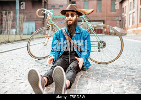 Ritratto di un barbuto hipster vestito elegantemente con il cappello e giacca seduta con retro bicicletta su strada sul background urbano Foto Stock