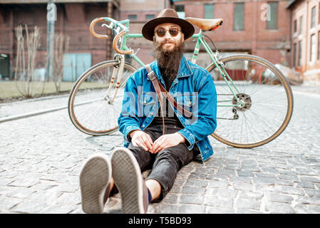 Ritratto di un barbuto hipster vestito elegantemente con il cappello e giacca seduta con retro bicicletta su strada sul background urbano Foto Stock